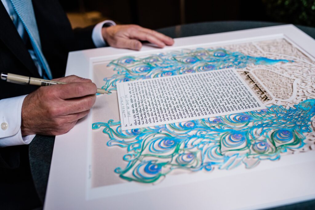 Signing the Peacock Ketubah papercut artwork with hand-painted watercolor peacock feathers and gold leaf calligraphy